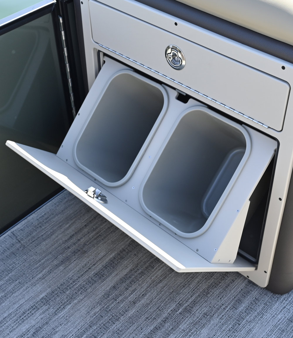 The lower door of a pontoon boat storage console is tilted open, revealing two integrated gray plastic trash bins for convenient waste disposal on deck.