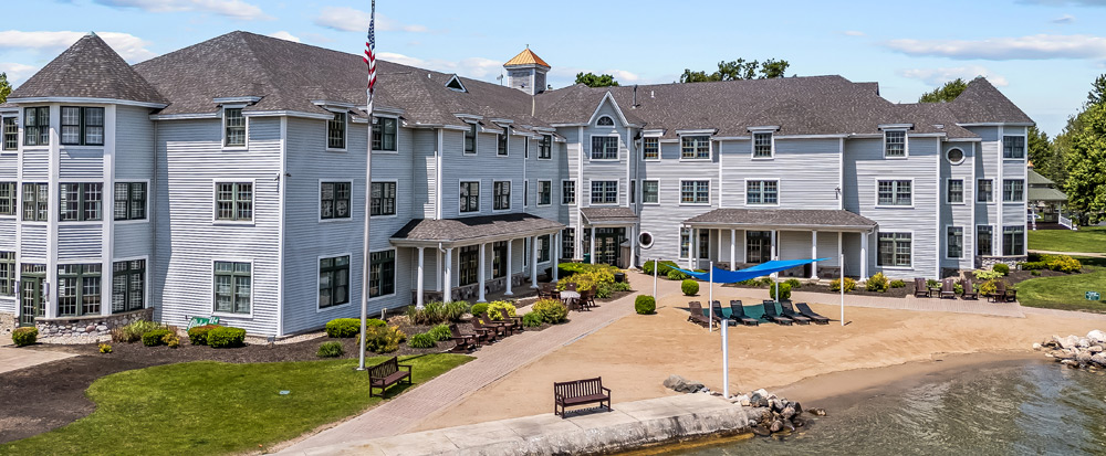 High-angle view of a large, gray three-story lakeside resort with a private sandy beach, lounge chairs, a blue sunshade, and a flag pole on the lawn.