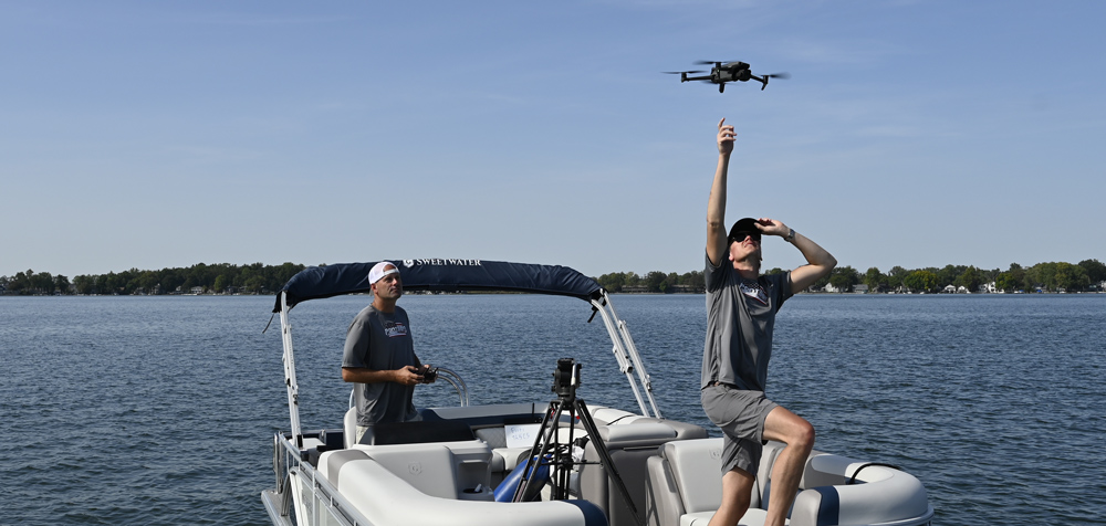 A man on a pontoon boat prepares to hand-launch a drone into the sky while a pilot operates the remote nearby on a clear, sunny day on the lake.