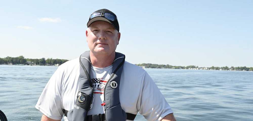 Portrait of a man wearing a gray performance shirt, a black "Pontoon" baseball cap, and a gray Mustang Survival inflatable life jacket on a boat.