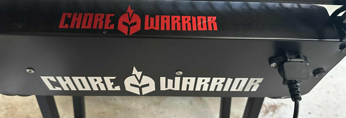Close-up of a black metal panel featuring the "CHORE WARRIOR" logo in red and white text. A black charging cord is plugged into a port on the right side of the panel.