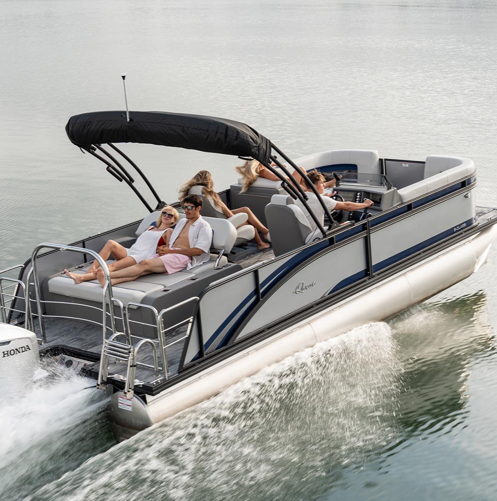 A light gray and blue Qwest pontoon boat cruises on water with five people on board. A man drives while two passengers lounge on the stern sun pad. The boat features a black bimini top and a Honda outboard motor creating a wake.