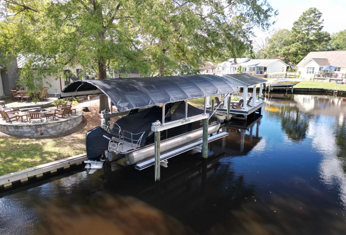 A pontoon boat with a black cover sits on a lift beneath a large, black, curved canopy with a silver aluminum frame. The boat and canopy are located at a residential waterfront dock with houses and trees in the background.