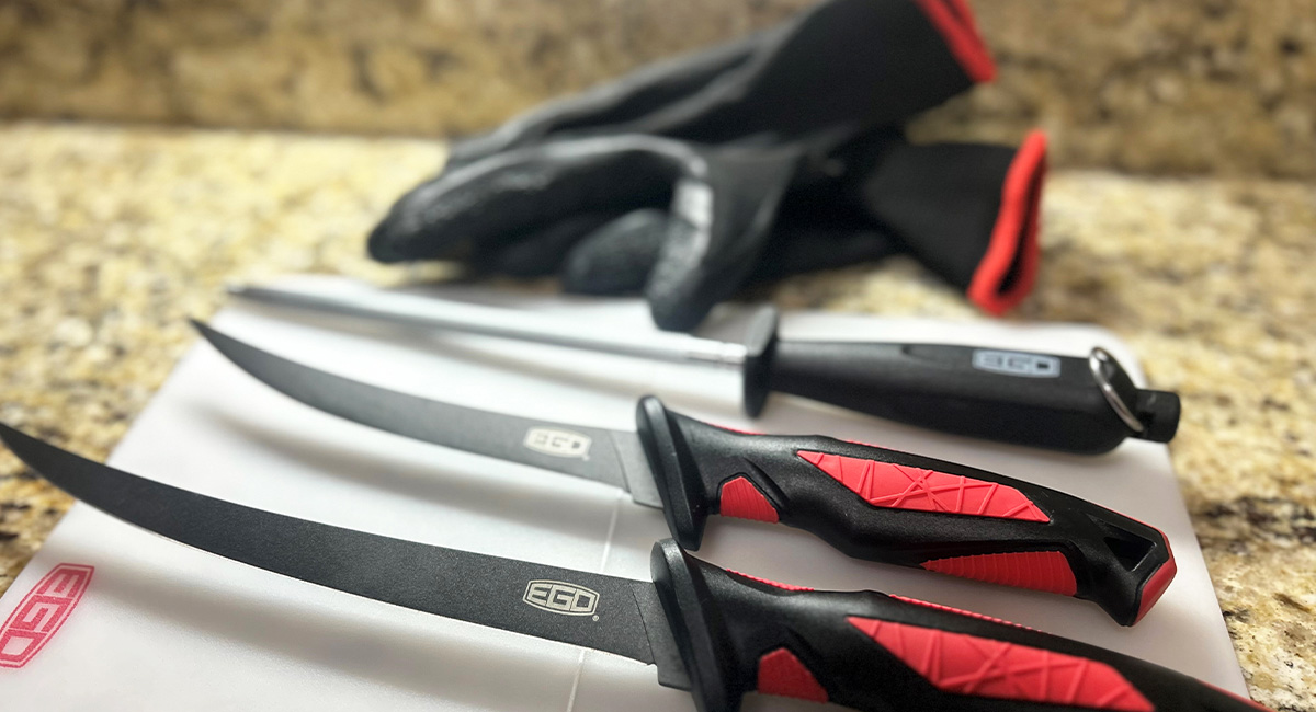 Two black fillet knives with red-textured handles, a sharpening steel, and black gloves with red trim are arranged on a white cutting board. The EGO logo is visible on the knife blades and the corner of the board.