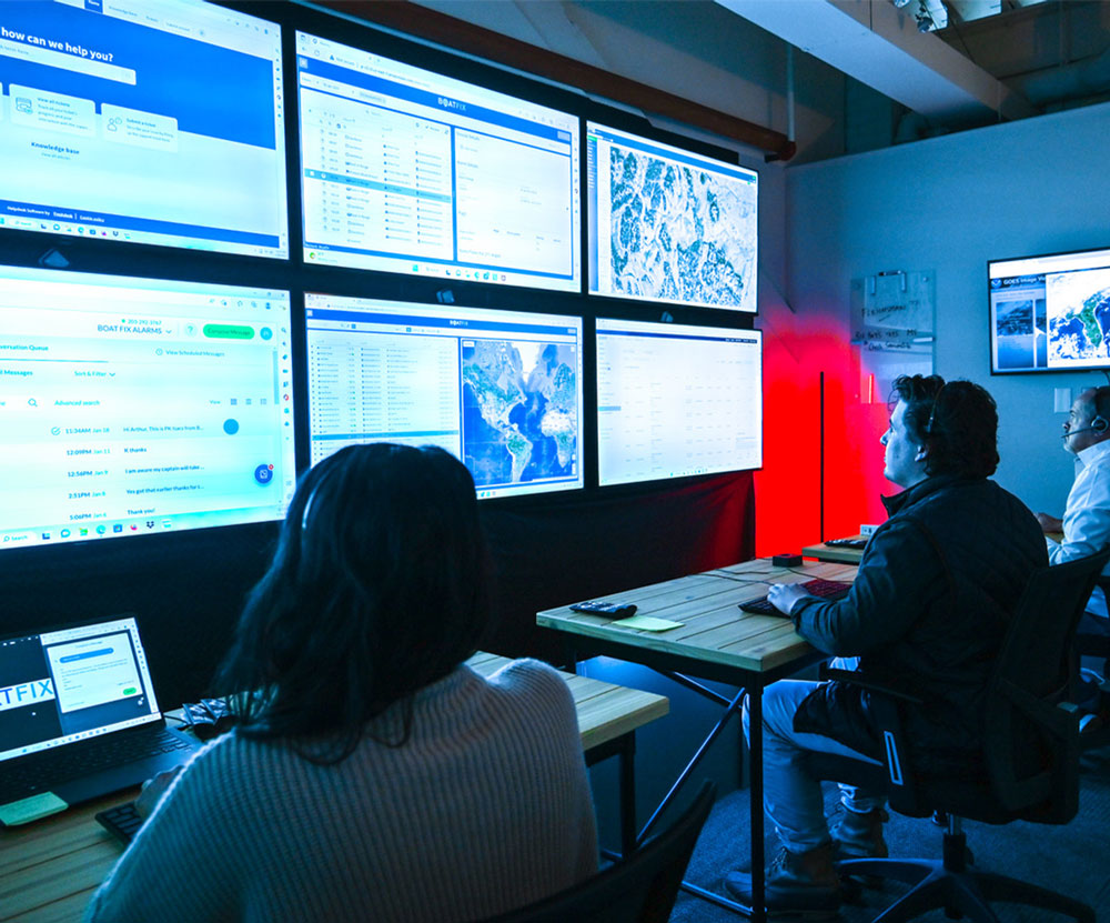 Three people wearing headsets sit at desks facing a wall of six large monitors. The screens display data dashboards and maps. The room is dimly lit by blue screen light and a red light in the corner.