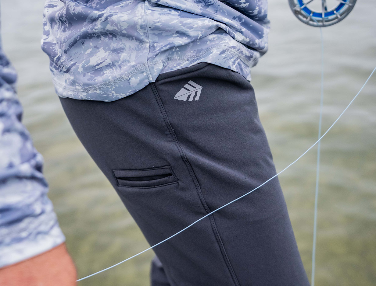 Close-up of dark gray joggers featuring a horizontal thigh pocket and a small logo near the hip. The wearer has a light blue camouflage shirt, with fishing line and a reel partially visible against a blurred water background.