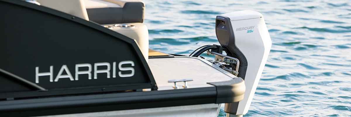 A close-up view of the stern of a black and tan Harris pontoon boat, featuring a white Mercury 35e Avator electric outboard motor mounted and tilted up.