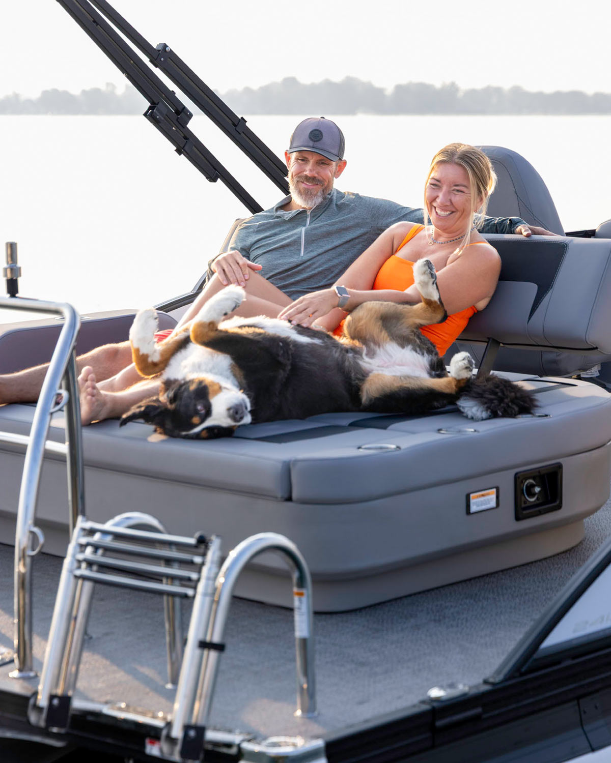 A couple smiles while relaxing on a spacious gray aft sunpad of a pontoon boat with a large dog lounging on its back.
