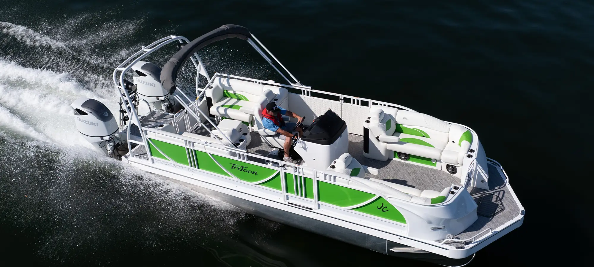 High-angle profile of a JC SportToon 26TT Twin HTH cruising on the water. Features lime green and white side panels, a black bimini top, and twin white Suzuki outboard engines. A driver is at the helm.