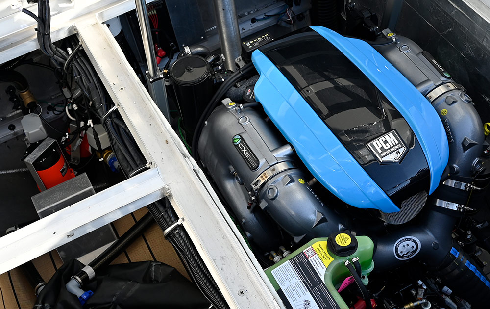 Interior view of the Montara EVO SL25 engine compartment containing a PCM marine engine with a light blue and black cover. Components include gray exhaust pipes, black hoses, and wiring bundles within the white internal hull.