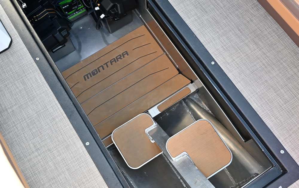 Close-up of an open floor locker on the Montara EVO SL25. The interior features tan faux-teak padding with "Montara" branding. A central metal support structure and battery components are visible within the compartment.