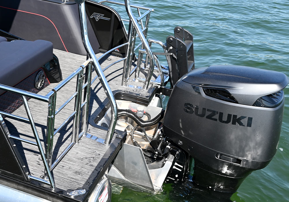 Rear view of the Qwest 24’ M-Class Lanai stern featuring a black Suzuki outboard motor. Includes a gray woven deck, stainless steel railings, a black boarding ladder, and a black storage compartment with red trim.