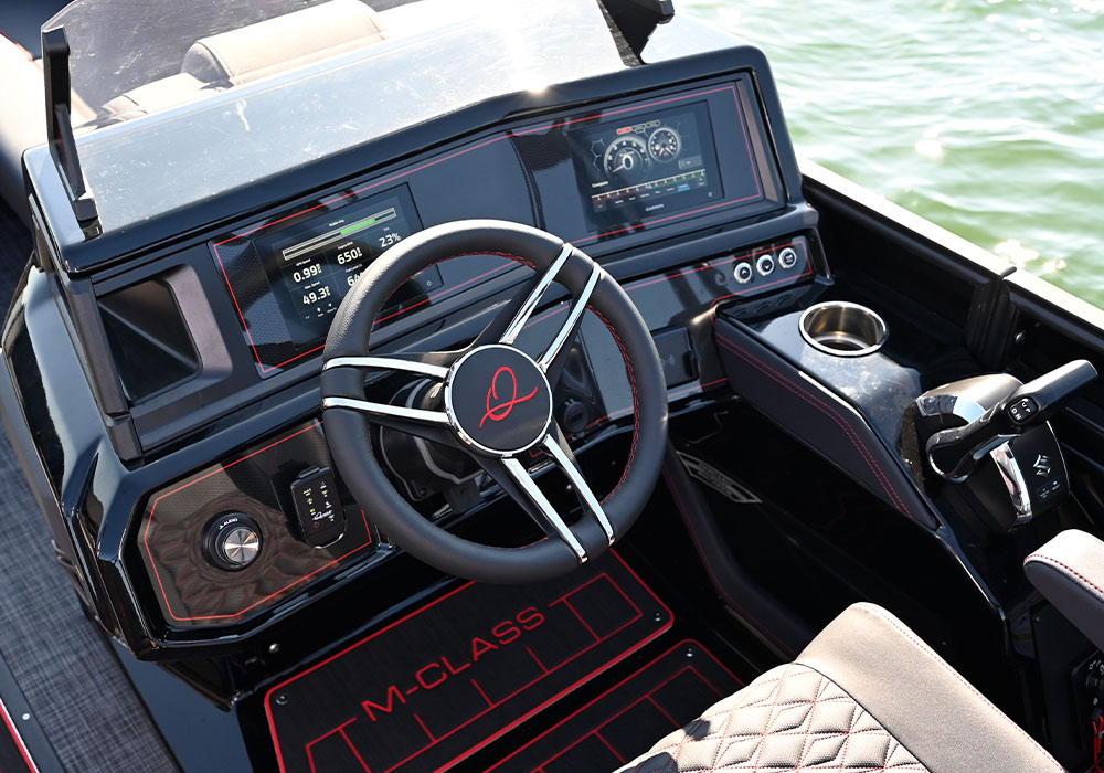The helm of a Qwest 24’ M-Class Lanai featuring a black dash with red trim and dual digital displays. Includes a steering wheel with a red "Q" logo, an "M-CLASS" branded floor mat, and diamond-stitched upholstery.