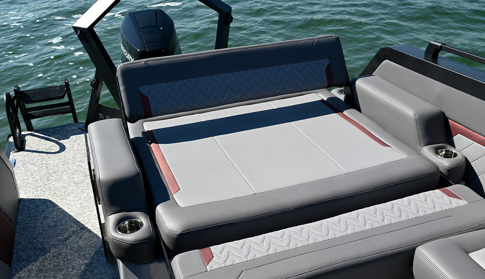 Close-up of the Revel RS25 rear lounge and sunpad area. Features gray upholstery with red accents and chevron-stitched patterns. Includes padded side bolsters with integrated stainless steel cup holders. A black Mercury motor is in the background.