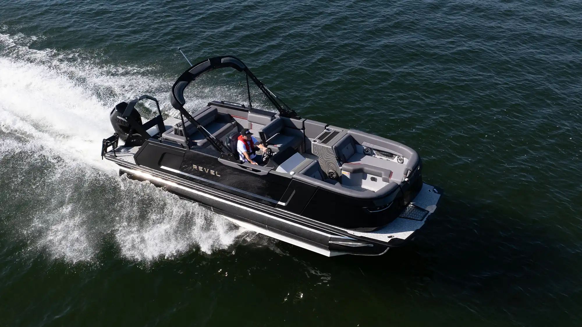 High-angle aerial view of a Revel RS25 pontoon boat cruising on the water. Features a black exterior with "REVEL" gold lettering, gray interior lounge seating, a black arch tower, and a single black Mercury outboard motor.