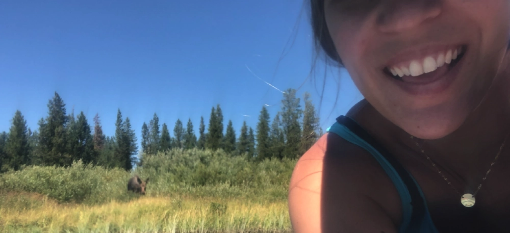 A close-up, partial selfie of a smiling woman in a blue tank top, with a grassy riverbank and a moose visible in the distant background under a clear sky