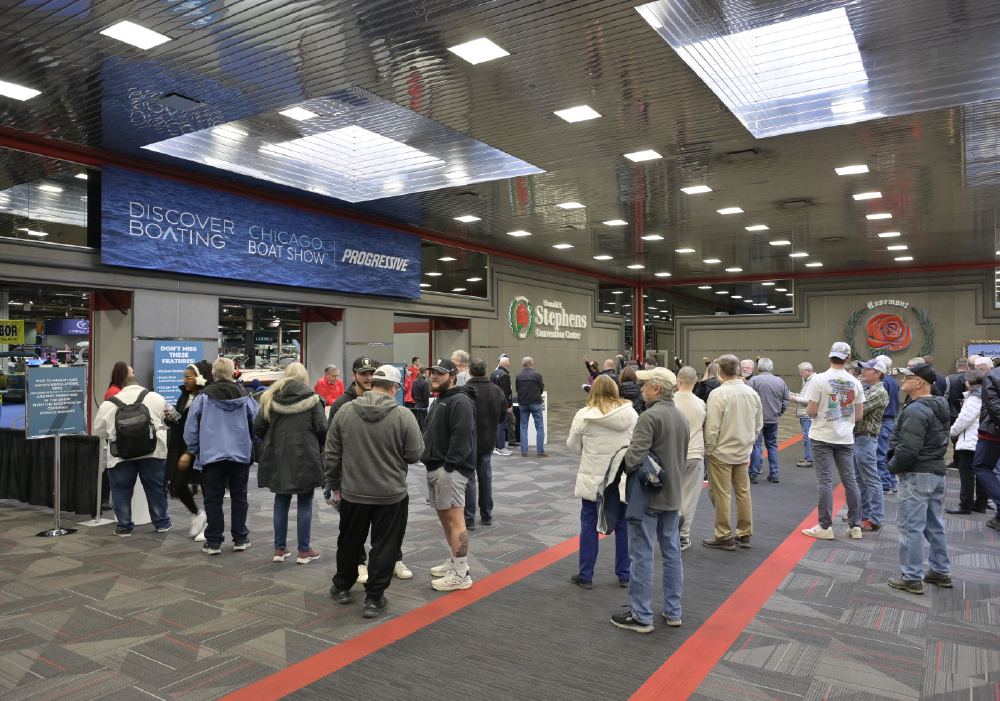 A wide interior view of the Donald E. Stephens Convention Center entrance; People are gathered in the lobby under a reflective metallic ceiling with square lights, and a blue banner above the entrance that reads the following logos DISCOVER BOATING | CHICAGO BOAT SHOW | PROGRESSIVE