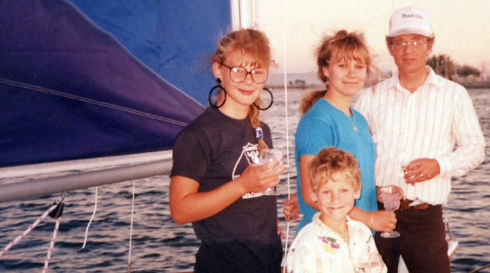 A vintage color photograph of a man and three individuals—two females and a young boy kid—standing on a dock next to a sailboat; They are all smiling and holding glasses of water