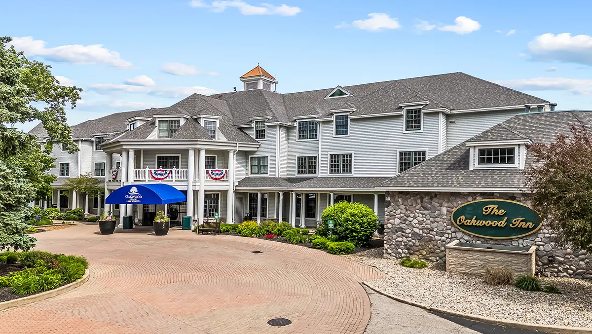 Exterior view of The Oakwood Inn resort at Lake Wawasee, featuring a large grey building with a stone entrance sign and a blue "Oakwood Resort" awning