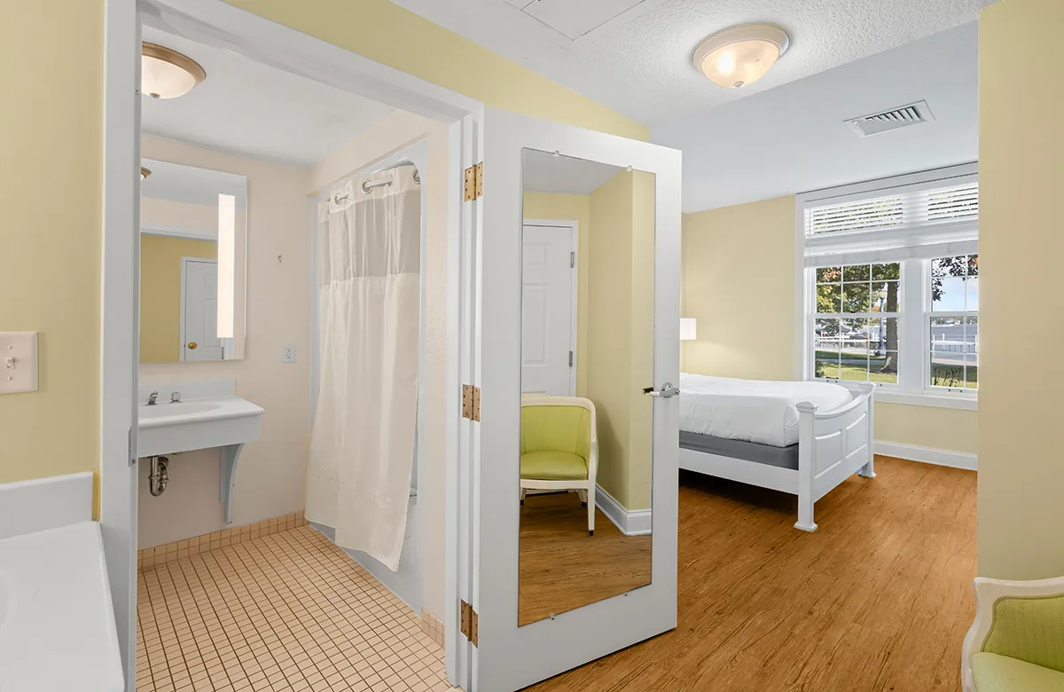 Interior view of a bright, modern guest room at Oakwood Resort featuring a white wooden bed, hardwood floors, and an en-suite bathroom with a vanity and walk-in shower
