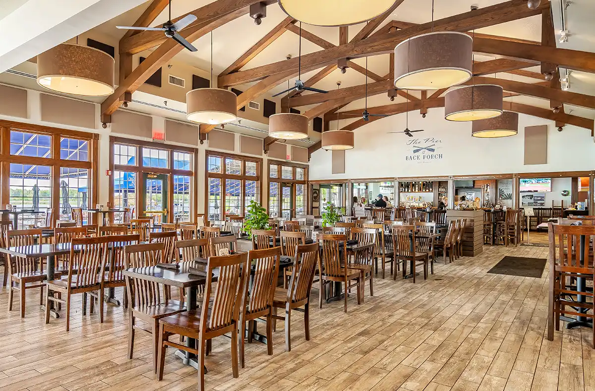 Interior view of "The Pier & Back Porch" restaurant at Oakwood Resort, featuring high vaulted ceilings with exposed wooden beams, rustic chandeliers, and ample wooden table seating.