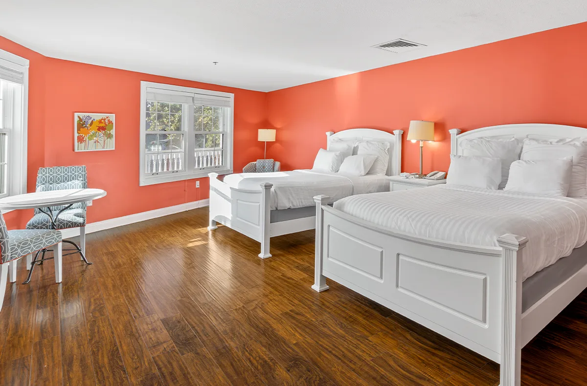 A bright, modern double-queen guest room at Oakwood Resort featuring two white wooden beds, polished hardwood floors, and a bold orange accent wall.