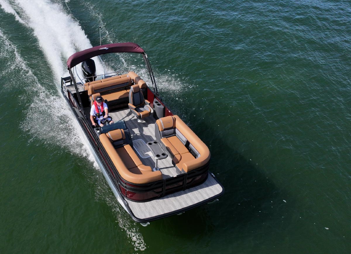 High-angle aerial view of a red and tan pontoon boat cruising across open green water, leaving a white wake behind it.