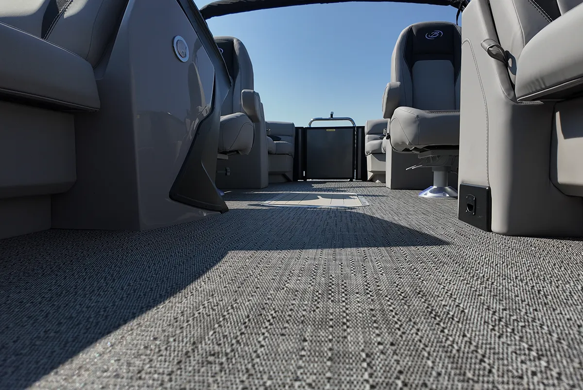 A low-angle interior view of a Barletta Aria pontoon boat, showcasing the durable grey woven marine flooring and high-back captain’s chairs under a clear blue sky.