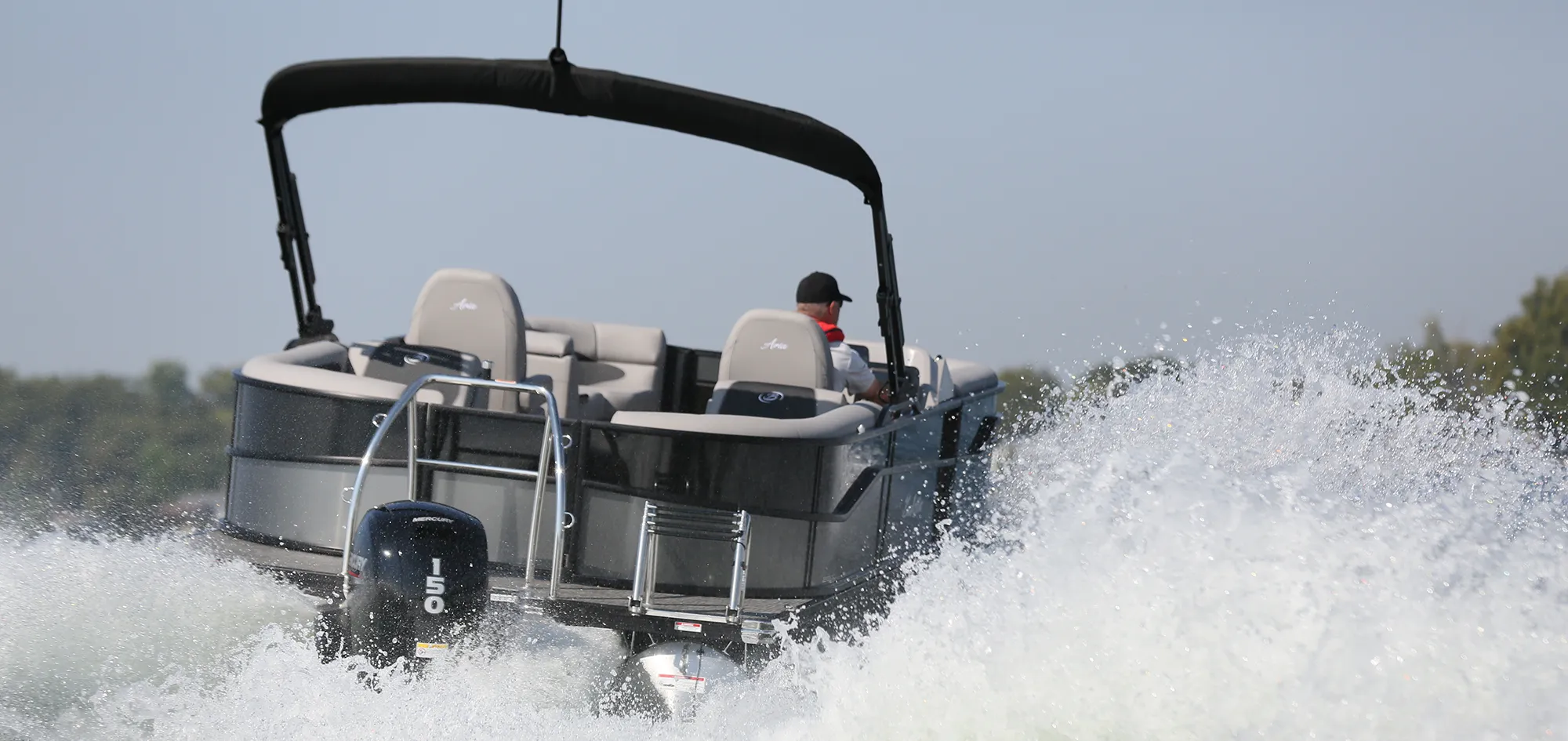 A Barletta Aria pontoon boat equipped with a 150-horsepower Mercury outboard motor cruising through choppy lake water, creating a large white spray.