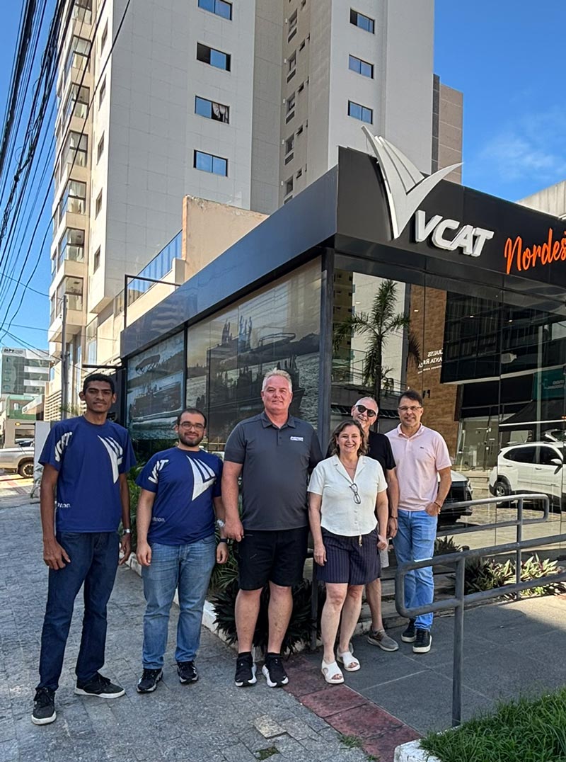 Five people stand on a sidewalk in front of a black VCAT Nordeste office. Two men wear blue branded t-shirts, and three others wear casual attire. A large white VCAT logo is visible on the building, with a tall apartment block in the background.