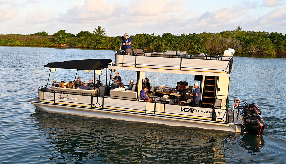 A large double-decker VCAT Alvorada 1100 pontoon boat on the water with many passengers. The boat features a main deck with a shaded seating area, a kitchen bar, and an upper sun deck. Two black Mercury Pro XS outboard motors are at the stern.