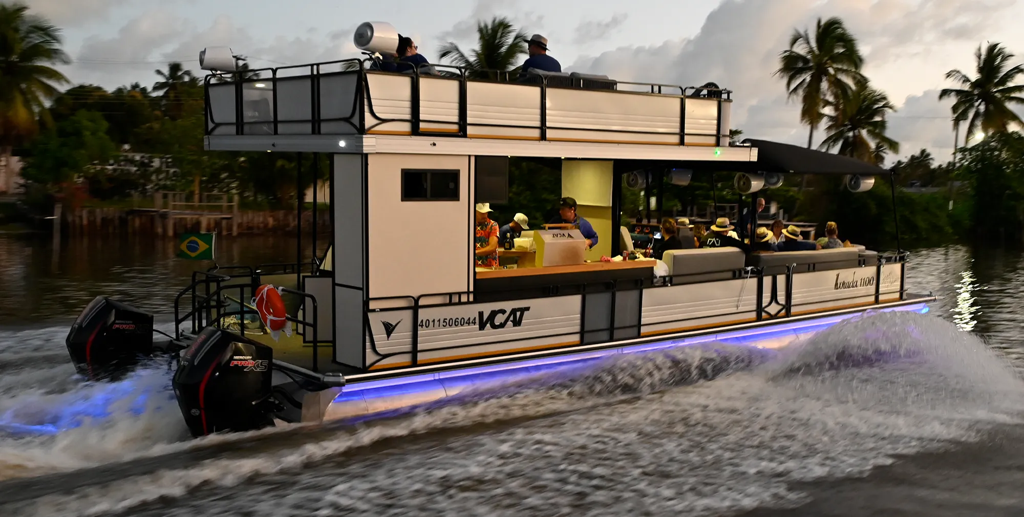 A VCAT Alvorada 1100 double-decker pontoon boat cruises at dusk with blue LED lighting along the pontoons. Passengers occupy the upper deck and a main-deck bar. Dual Mercury outboard motors power the vessel, creating a white wake.