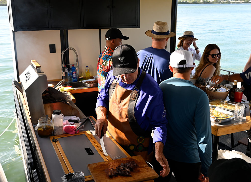 A chef in a purple shirt and leather apron slices grilled meat on a wooden board in a boat's outdoor kitchen. In the background, passengers stand near a food counter overlooking the water.