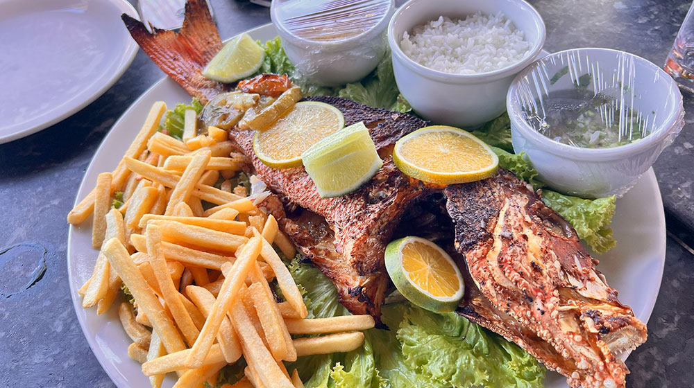 A whole grilled fish garnished with lemon and lime slices is served on a large white platter alongside French fries, a bowl of white rice, and small containers of sauces on a bed of green lettuce.