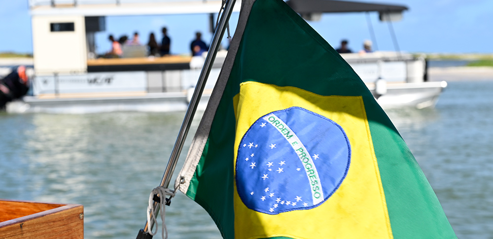 A close-up of a green and yellow Brazilian flag attached to a boat's railing. In the blurred background, a white and black double-decker VCAT pontoon boat moves across the water.