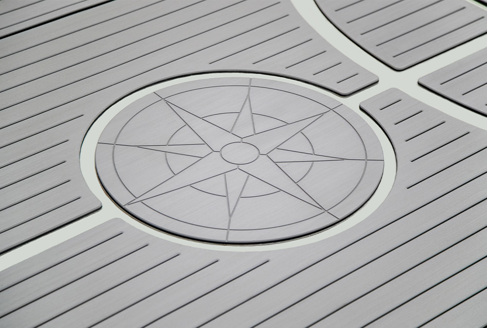 Close-up of SeaSole gray composite decking featuring a circular compass rose inlay. The slip-resistant surface is designed with parallel grooves and a peel-and-stick installation system.