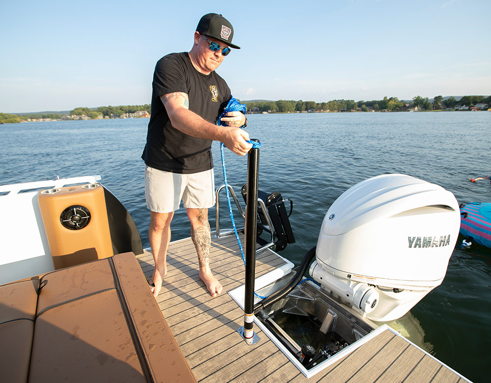 A man on a boat deck secures a blue rope to a black tow pylon next to a large white Yamaha outboard motor. The boat is on a calm lake with a tree-lined shoreline in the distance.