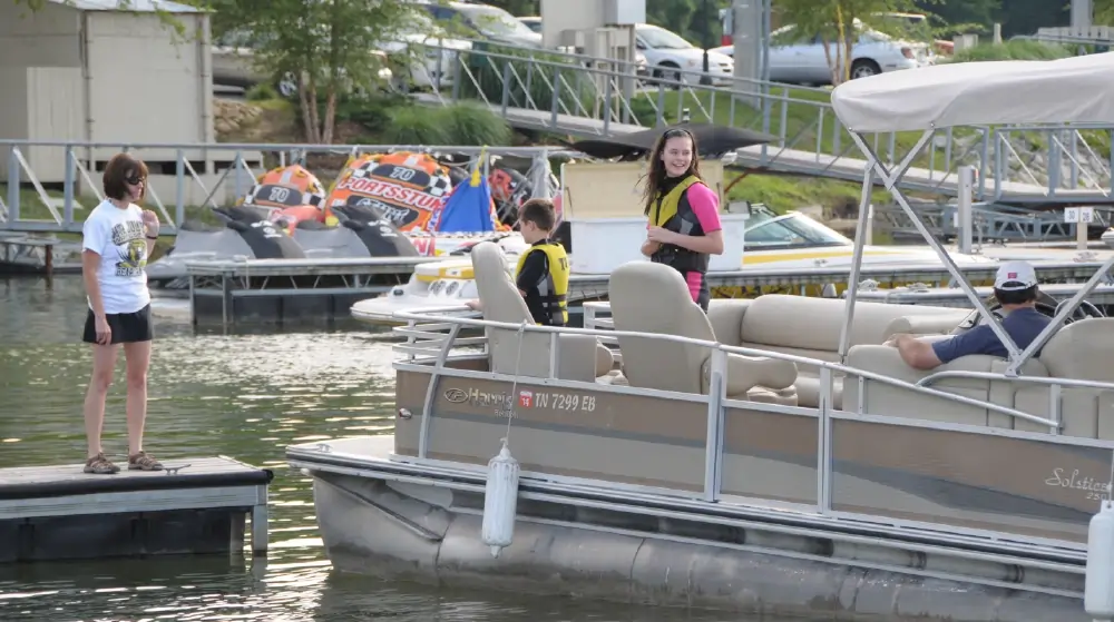 A young girl in a pink swimsuit, a young boy in a black swimsuit, and a woman standing on a nearby dock while a man sits at the helm of the boat