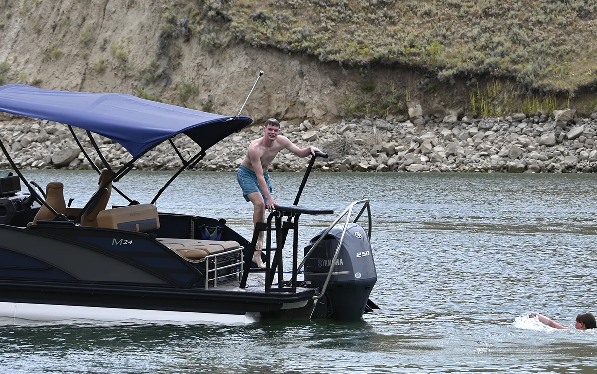 A young man climbs onto the stern of a Bennington M24 pontoon boat next to a Yamaha 250 outboard motor, with another swimmer visible in the water