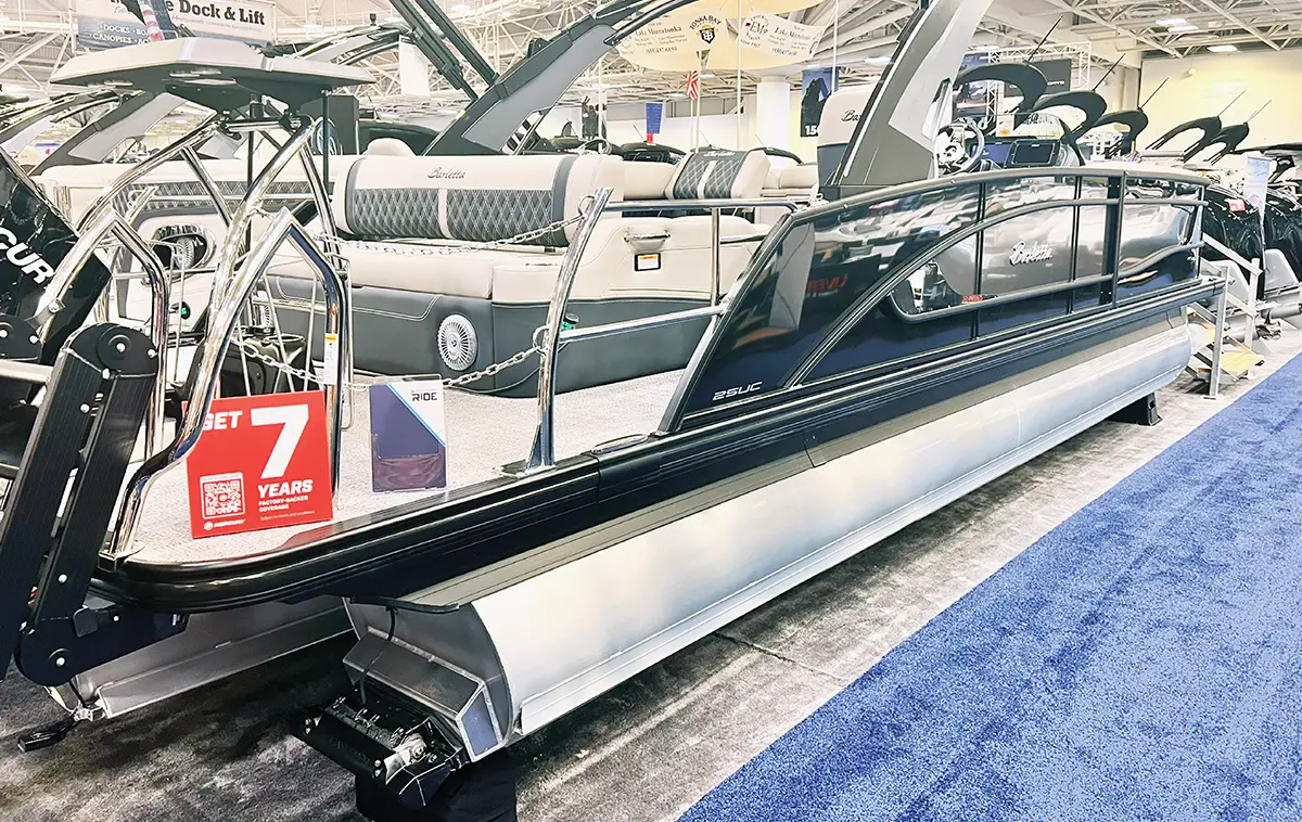 Profile view of a luxury Barletta pontoon boat on display at an indoor boat show, featuring sleek black rails, light grey upholstery, and a "7 Years Coverage" promotional sign.