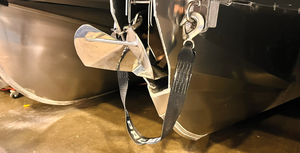 Close-up of a polished silver anchor stowed in a specialized housing on a pontoon boat hull, secured with a black "PREMIER" branded heavy-duty nylon safety strap and hook.