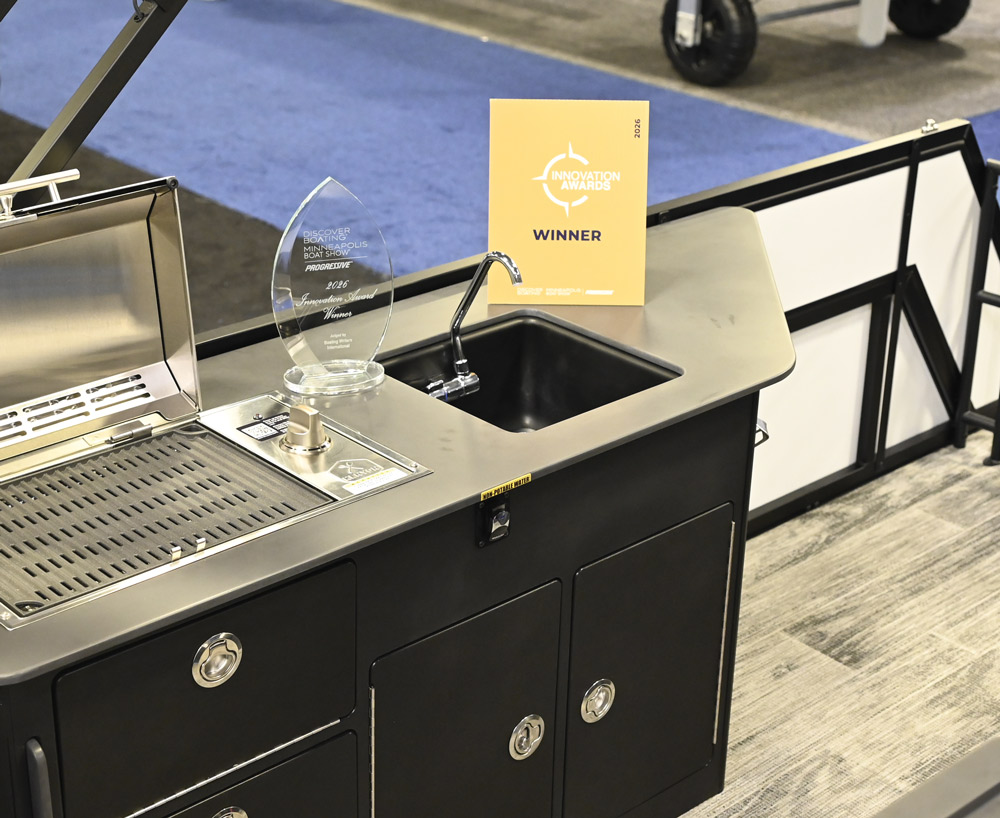 A black outdoor kitchen unit on a boat features a stainless steel grill, a small sink with a chrome faucet, and a 2026 Minneapolis Innovation Award trophy resting on the counter.
