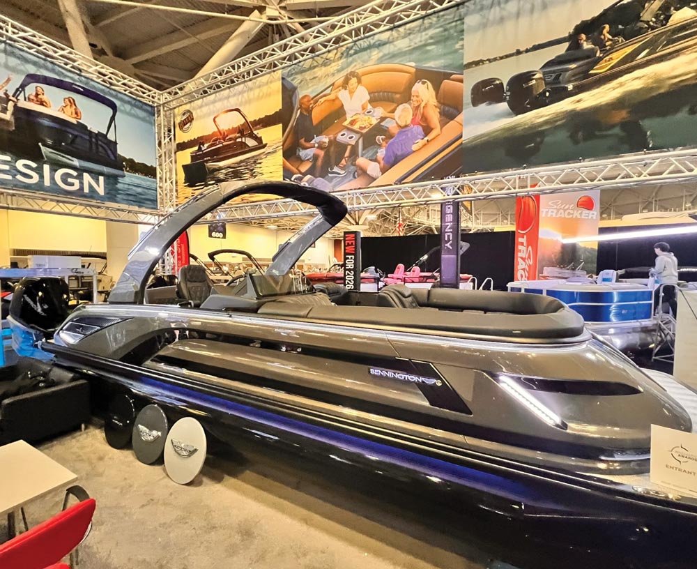 A sleek, modern Bennington pontoon boat in metallic grey with a large arch and LED accent lighting is displayed under bright rafters at a large indoor convention center.