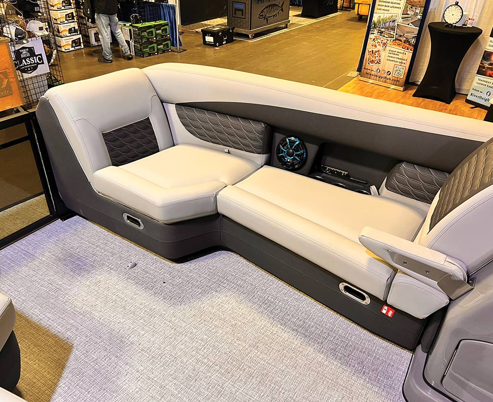 Interior view of a pontoon boat featuring a light grey L-shaped lounge seat with dark grey quilted accents, integrated blue LED speakers, and woven vinyl flooring.