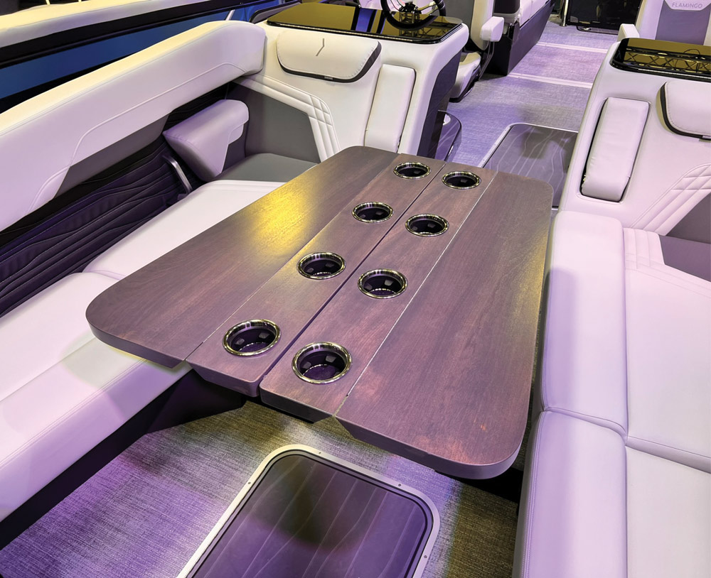 A dark wood-grain table with eight integrated stainless steel cup holders is positioned between light-colored lounge seats inside a luxury pontoon boat.