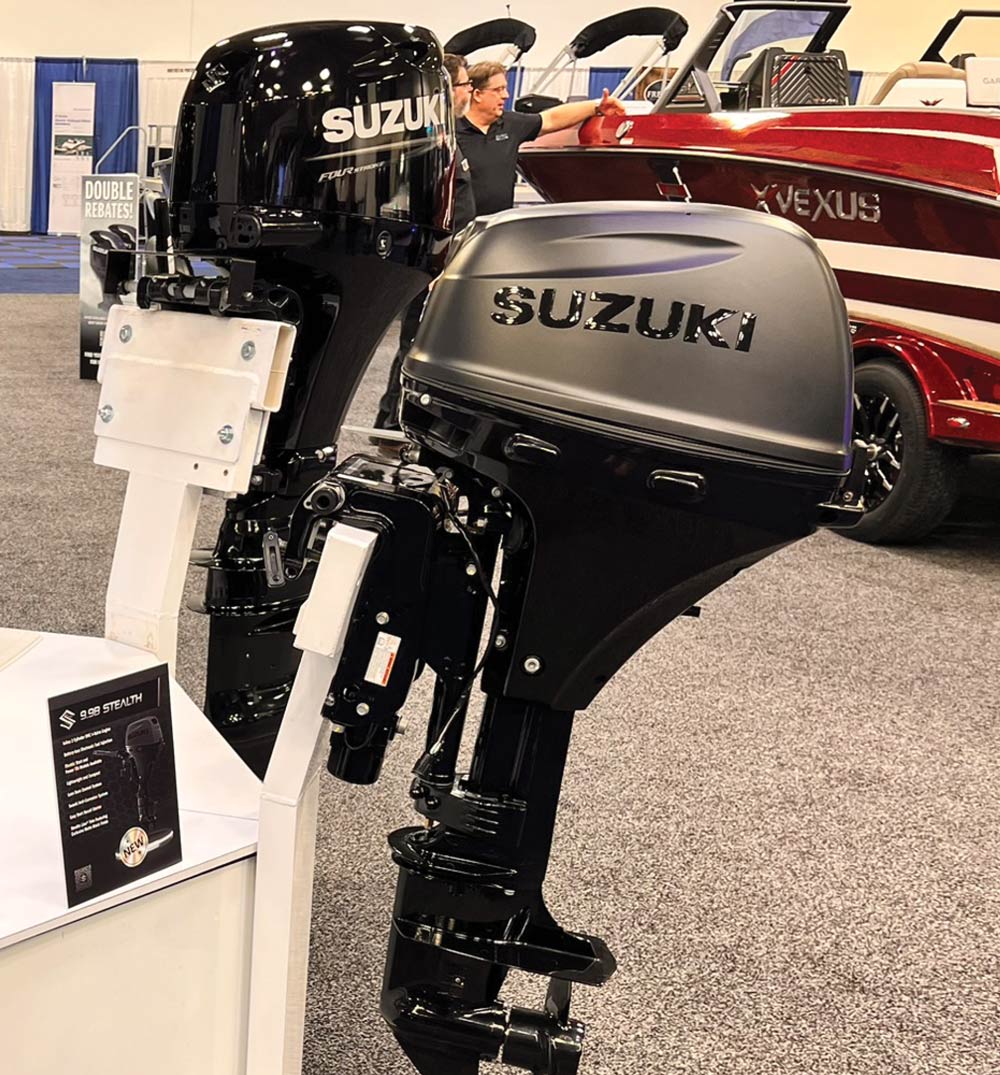 Two Suzuki outboard motors, one in gloss black and one in matte "Stealth" grey, are mounted on display stands at a boat show next to a promotional information card.