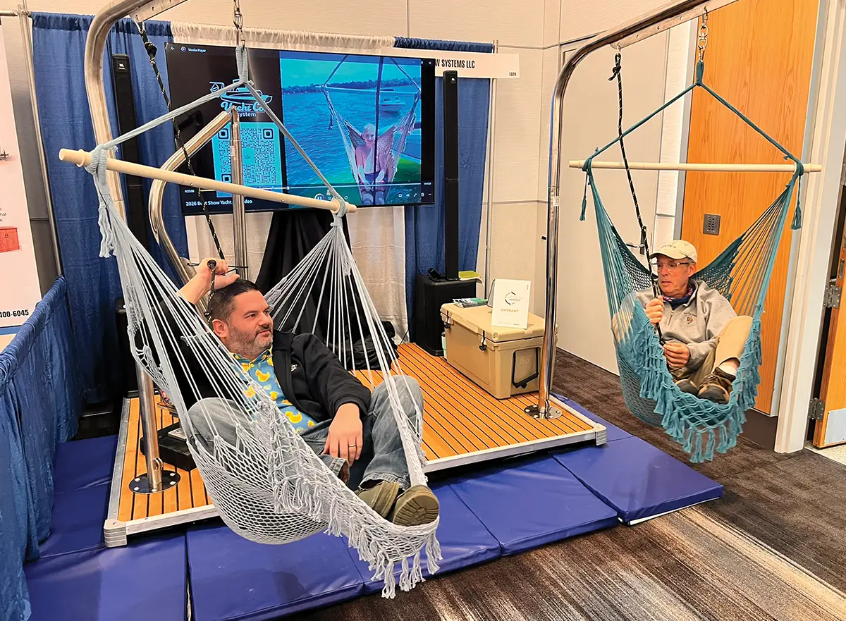 Two men test out hanging hammock chairs suspended from stainless steel frames at a boat show exhibit. Behind them, a television screen displays a video of the hammocks in use on the water.