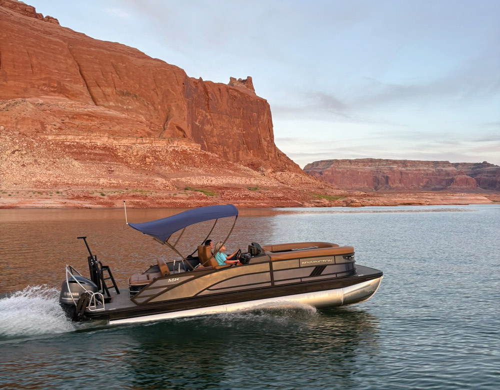 Pontoon boat speeding across water near red sandstone cliffs under a blue sky.