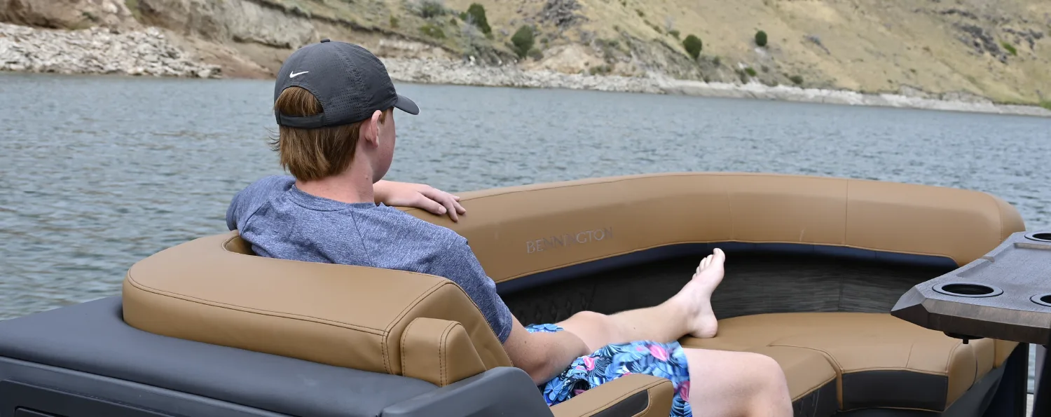 A passenger relaxes on the tan leather upholstery of a Bennington pontoon boat, looking out at the water and a hilly shoreline