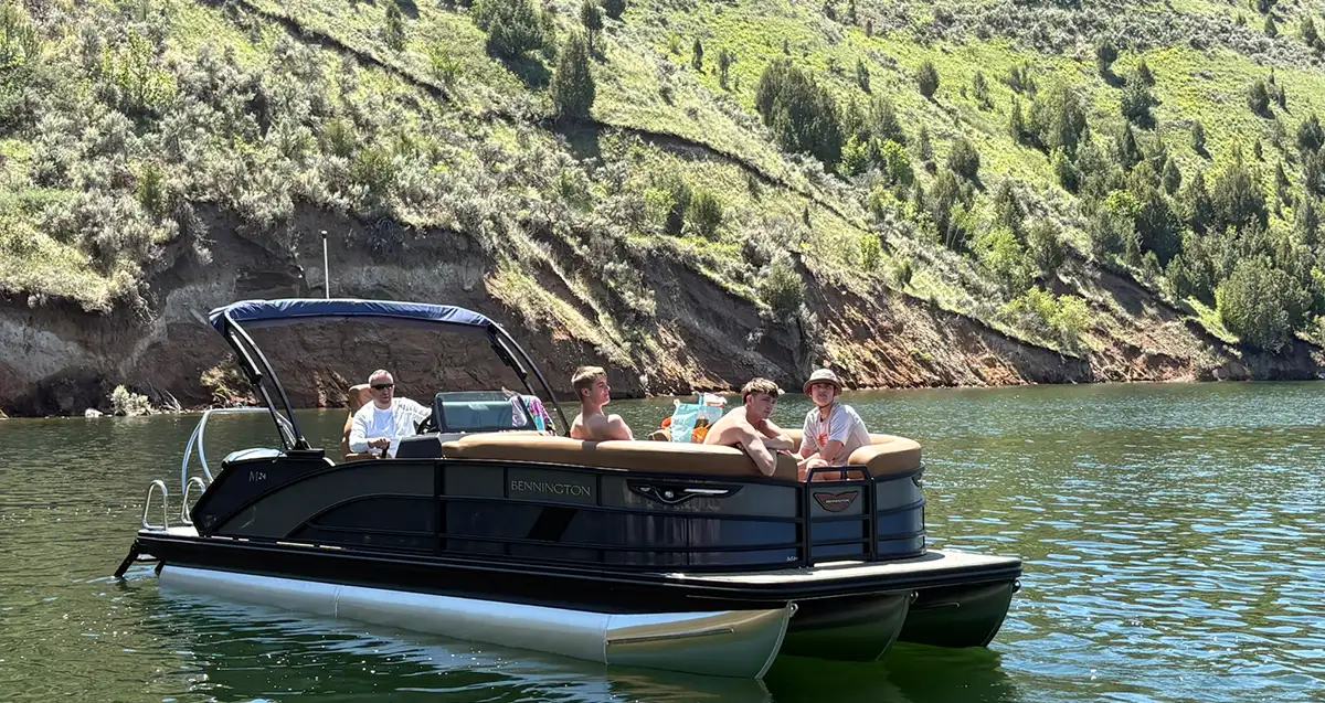 A black and tan Bennington tritoon boat with four passengers on board, floating on a calm lake in front of a steep, sunlit hillside